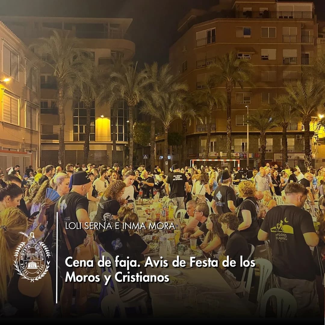 Multitud de personas cenando en mesas largas al aire libre durante una noche festiva en una plaza urbana con palmeras y edificios de fondo.