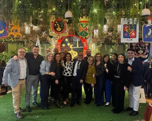 Grupo de personas posando sonrientes en un salón decorado con plantas colgantes, luces y escudos heráldicos sobre suelo de césped artificial.