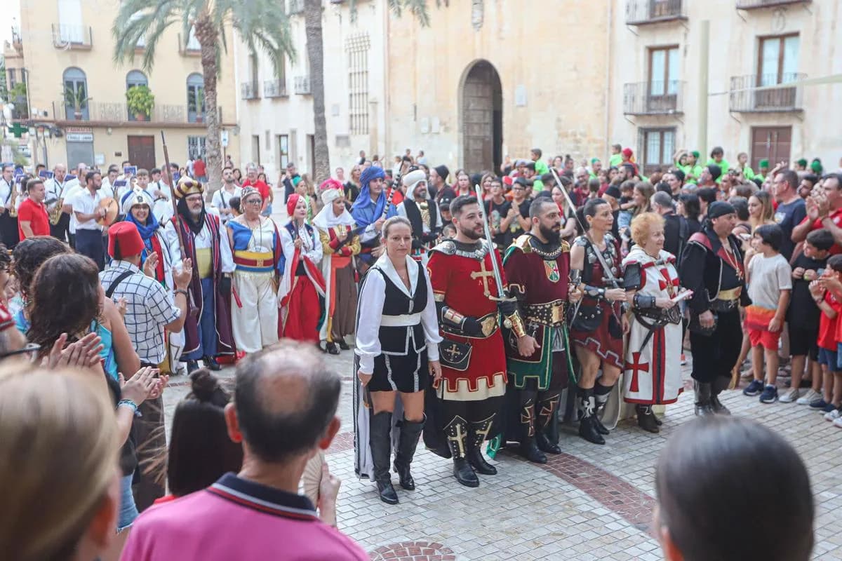 Personas con trajes tradicionales de Moros y Cristianos desfilan por una plaza ante un público que los observa y aplaude.