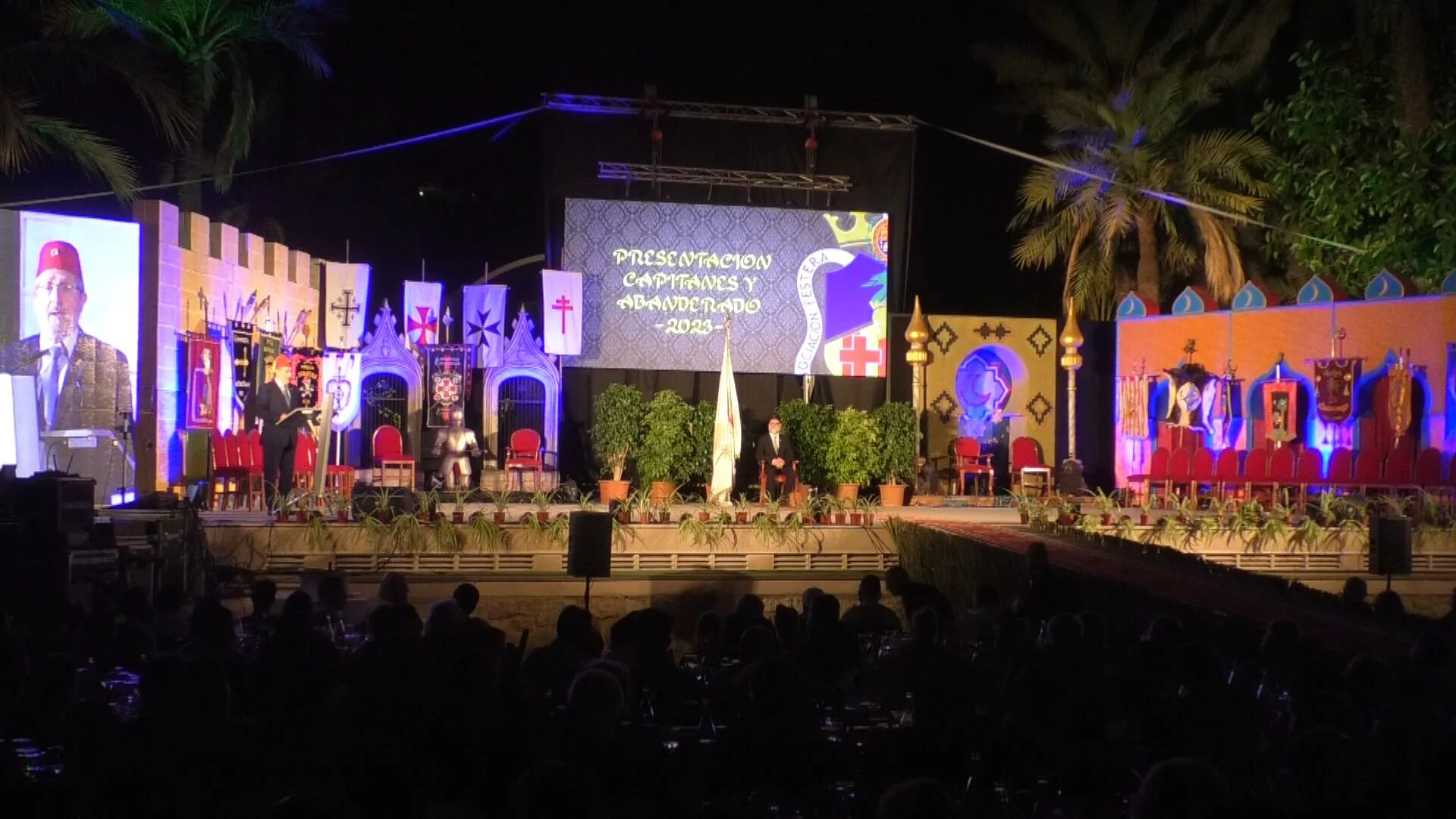 Escenario nocturno decorado con elementos cristianos y árabes para un acto de presentación frente a un público sentado.